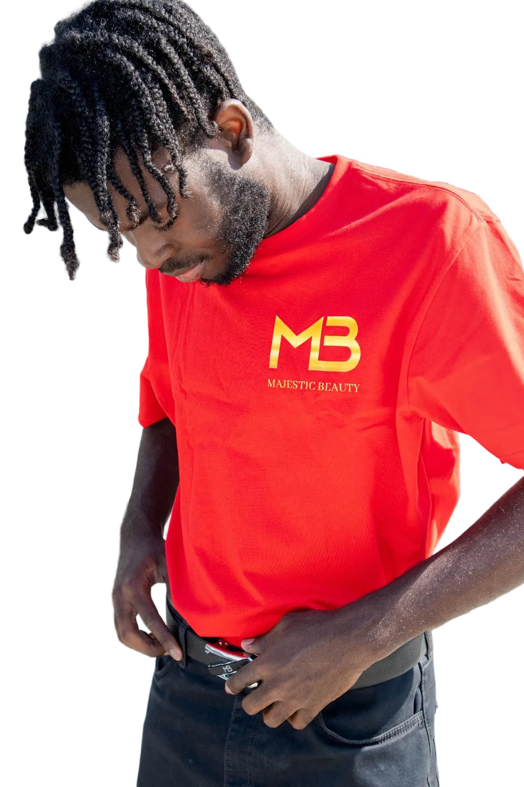 A man wearing a bright red crewneck t-shirt with a yellow 'MB MAESTIC BEAUTY' logo on the chest, standing outdoors next to a stone monument.